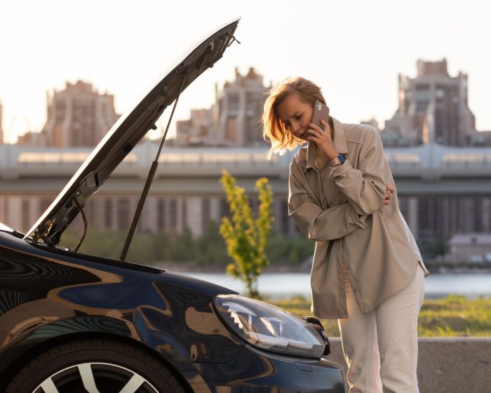 Woman driver calling for help assistance looking at broken car with opened hood, talking on mobile